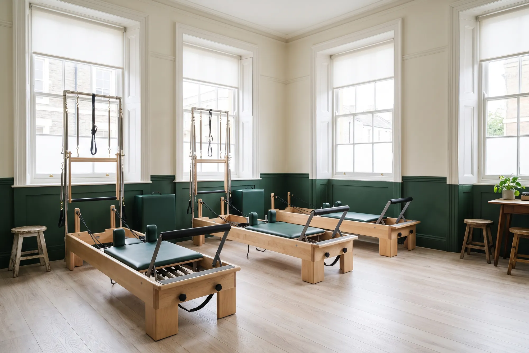 Three wooden Pilates reformers with green upholstery in a bright studio with tall windows.