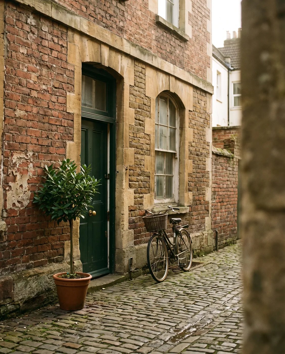 A green front door in an old brick and stone building on a cobbled lane, with a bay tree in a pot and a bicycle against the wall.